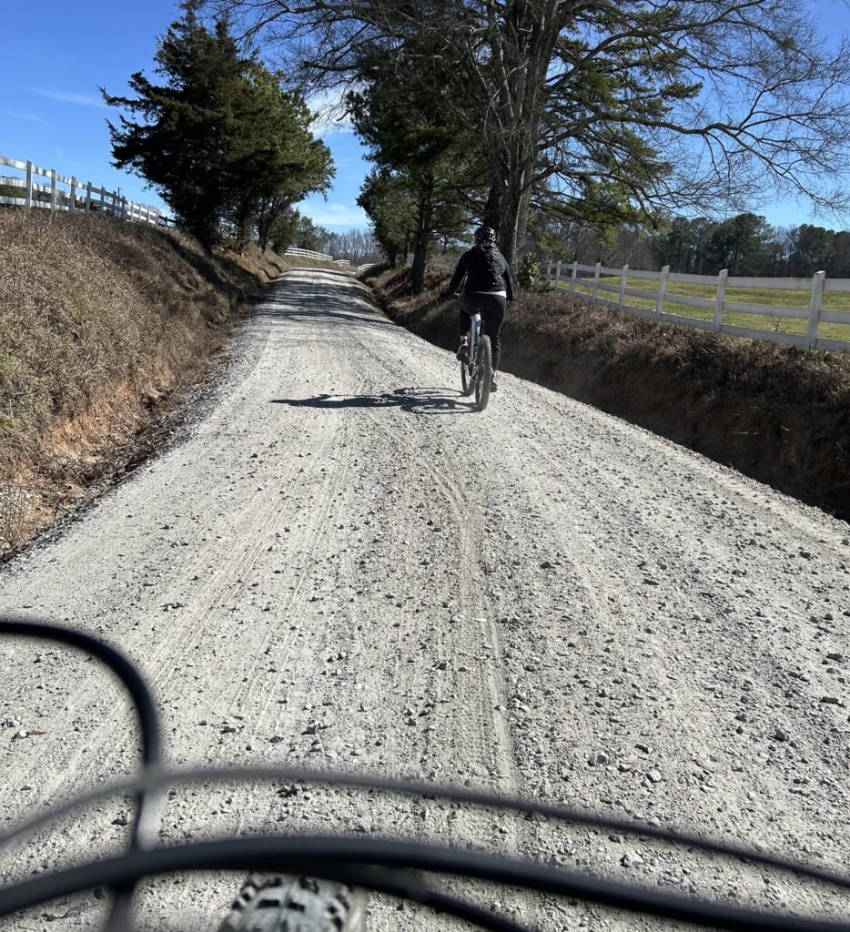 An eMTB biker is riding on a gravel road through farmland, from the perspective of a biker behind them.