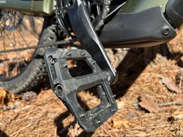 A closeup of a black pedal on a green eMTB. Dirt trail is the backdrop.